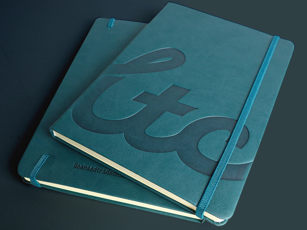 Two teal notebooks with elastic bands are stacked, with the top one featuring a bold, embossed design. Both have a smooth, matte cover and rounded corners, set against a dark background.