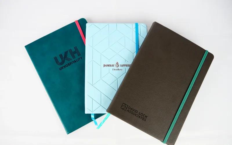 Three branded notebooks in teal, light blue, and brown, featuring logos for UKH, Bombay Sapphire, and David Lock Associates.