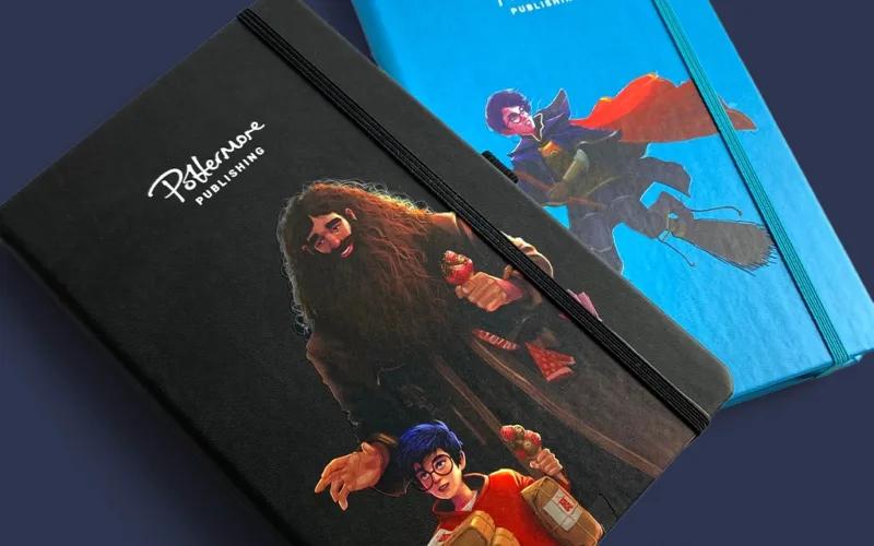 Two hardcover notebooks with elastic bands are shown. The black notebook features illustrated characters resembling a wizard holding an umbrella and a boy with glasses. The blue notebook shows a boy riding a broomstick.