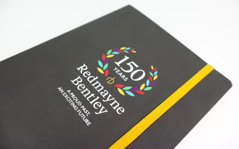 A black notebook with a yellow elastic band features “150 Years Redmayne Bentley” and a colorful laurel wreath graphic, along with the text “A Proud Past, An Exciting Future” on the cover.