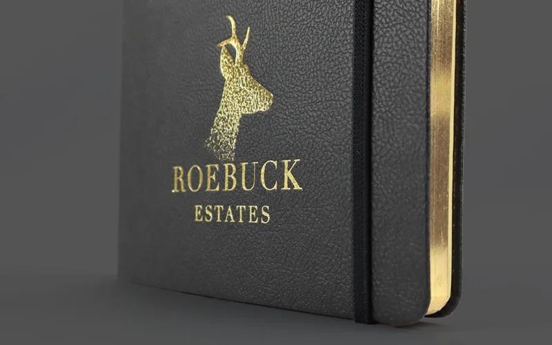A black notebook with gold-edged pages features a gold embossed deer head and the words ROEBUCK ESTATES on the cover. An elastic band is attached to the side.