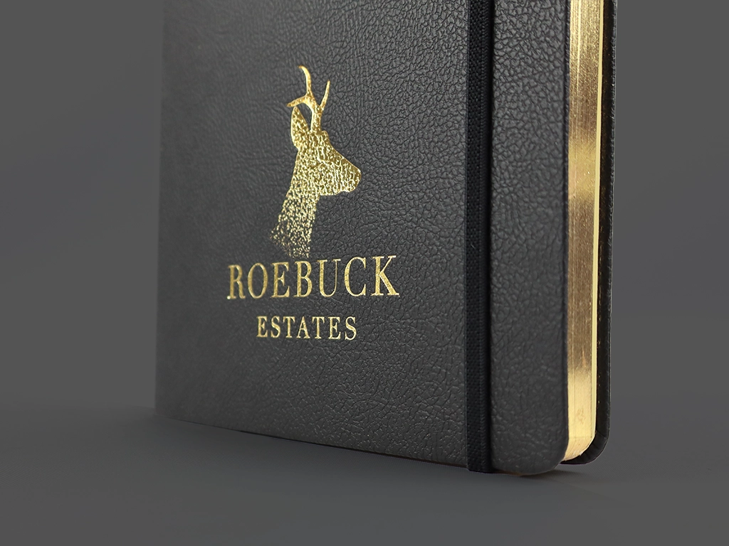 A black notebook with gold-edged pages features a gold embossed deer head and the words ROEBUCK ESTATES on the cover. An elastic band is attached to the side.