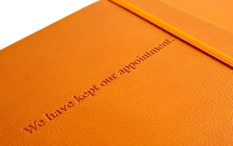 An orange textured notebook with an elastic band and embossed text on the cover that reads, We have Kept our appointment.