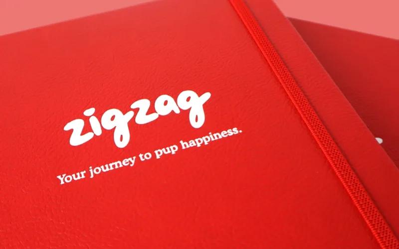A close-up of a red notebook with an elastic band, featuring the white text zigzag and the slogan Your journey to pup happiness. on the cover. The background is a matching red shade.