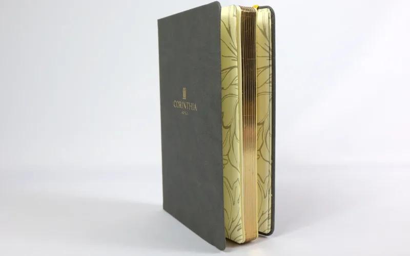 A hardcover book with a dark gray cover and gold embossed text stands partially open, revealing gold-edged pages with a floral pattern design. The background is plain white.