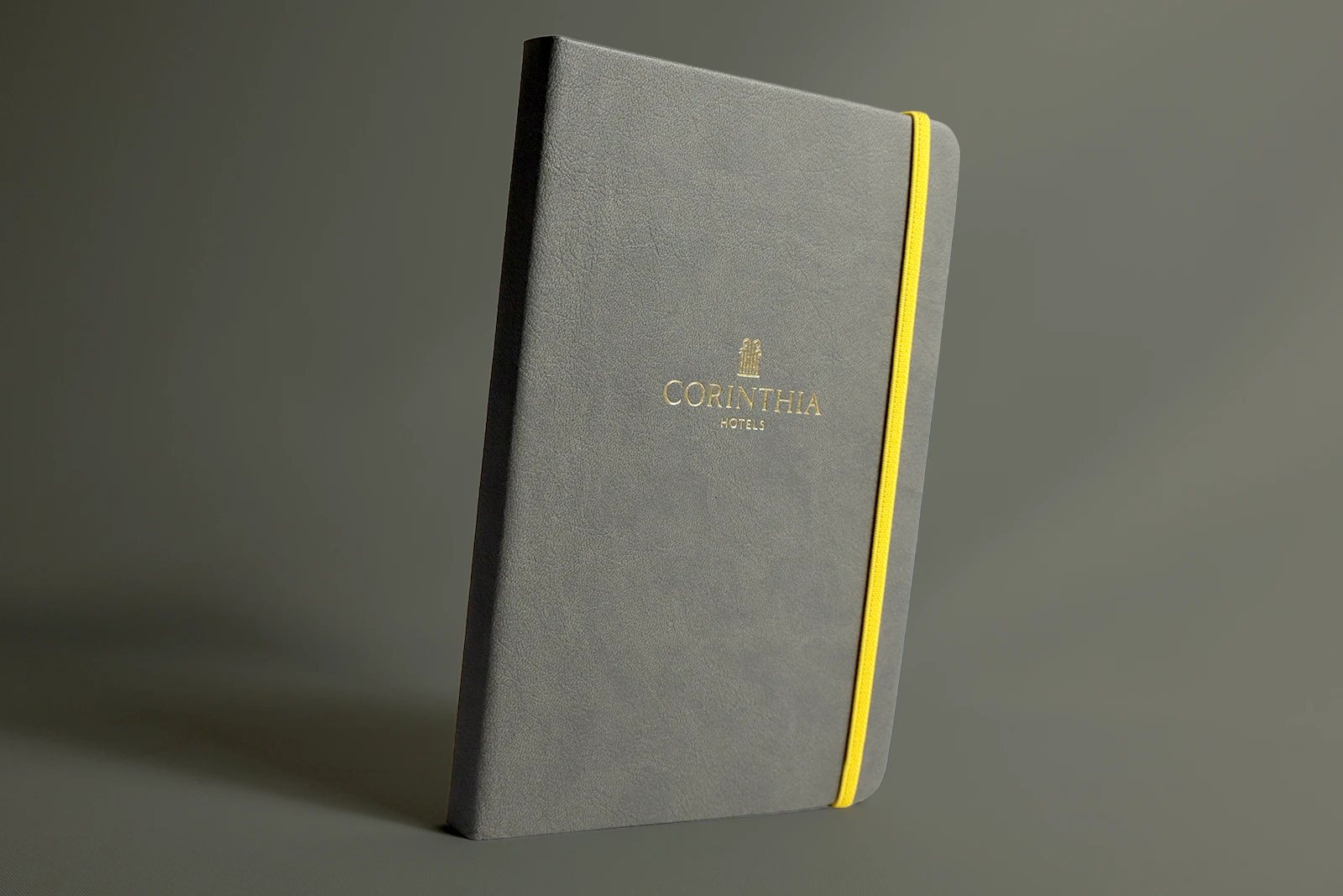 Front view of Corinthia branded notebook. It is dark grey with a gold foil logo and yellow elastic closure band.