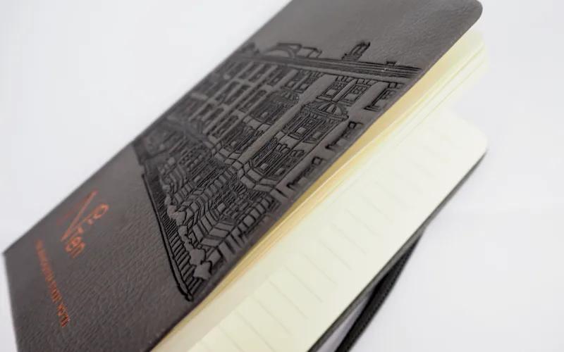 A black leather notebook with an embossed building design on the cover, open to reveal lined cream-colored pages.