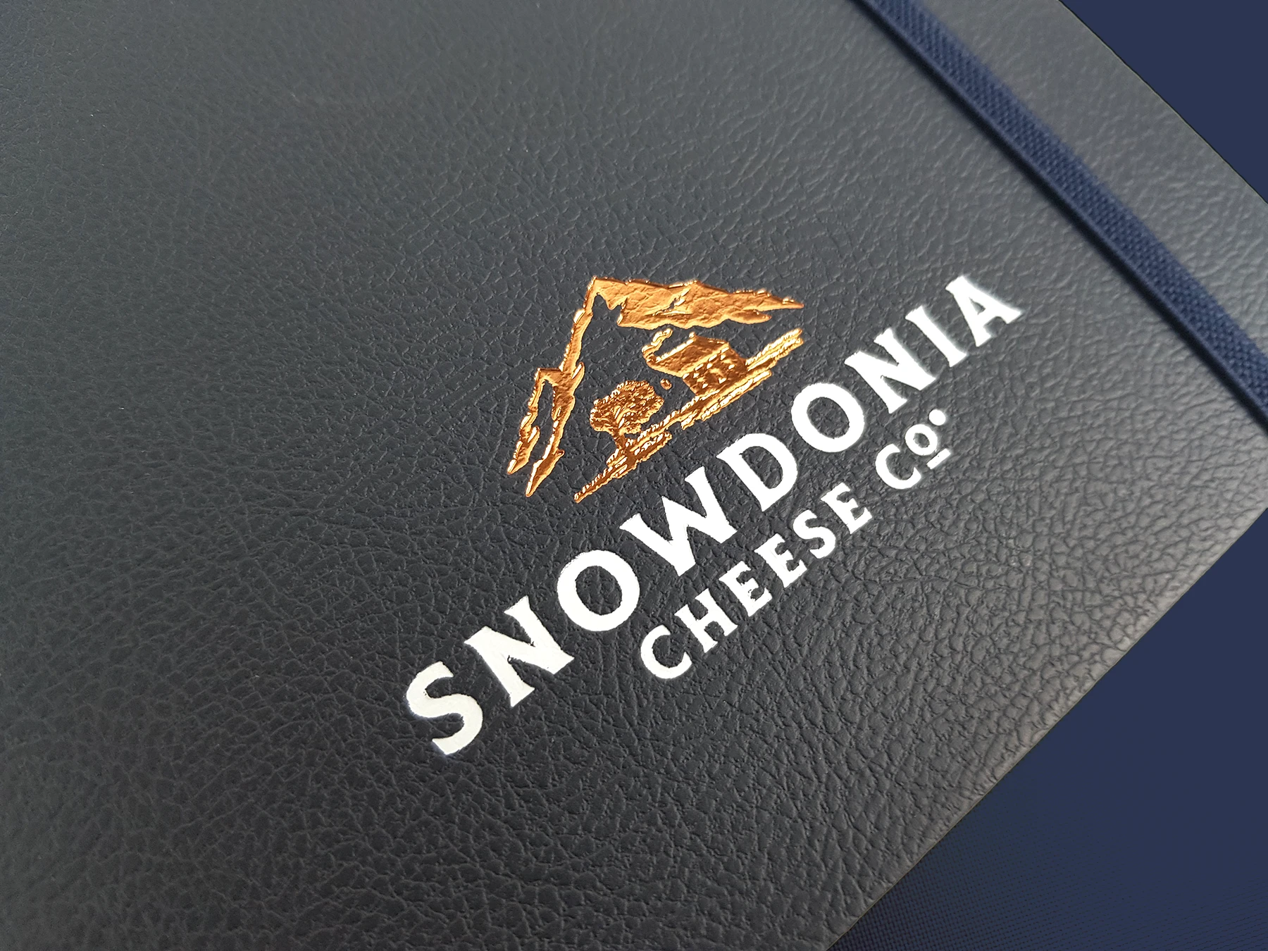 Close up of Snowdonia cheese co. branded notebook in navy blue, with a copper and white foil printed logo in the bottom center.